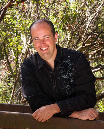 The image features a man standing outdoors, leaning on a fence with one hand while the other is casually placed in his pocket. He is smiling and looking directly at the camera.