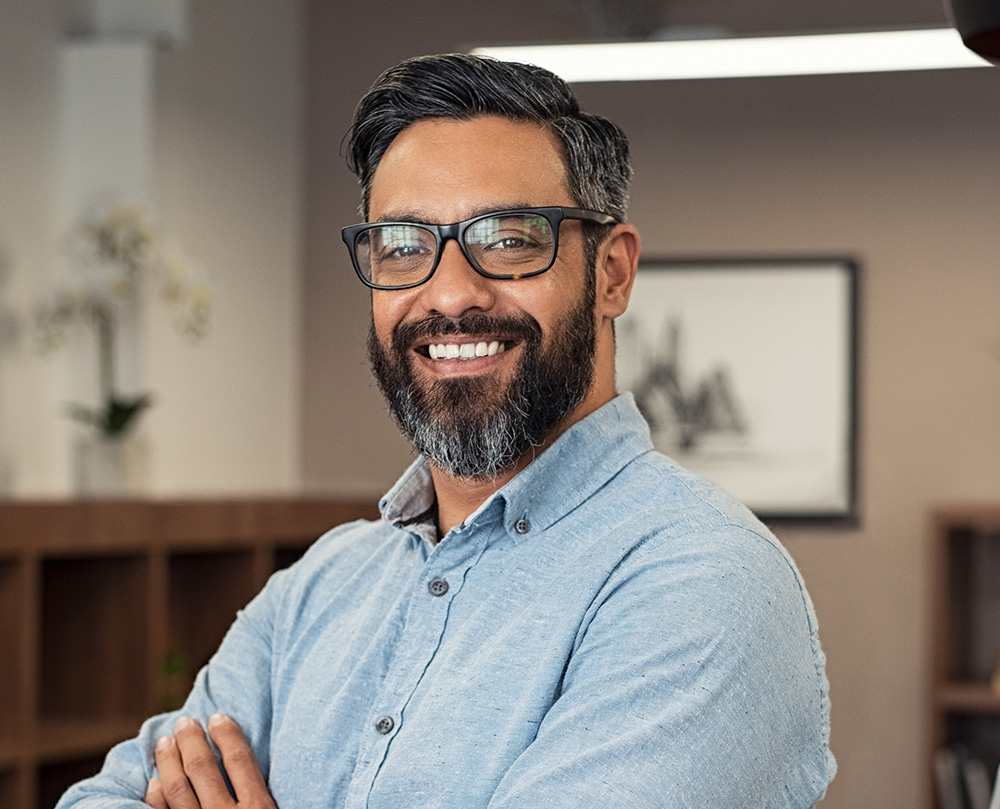 The image shows a man with a beard and glasses, wearing a blue shirt, standing in an office environment. He has his arms crossed and is smiling at the camera.