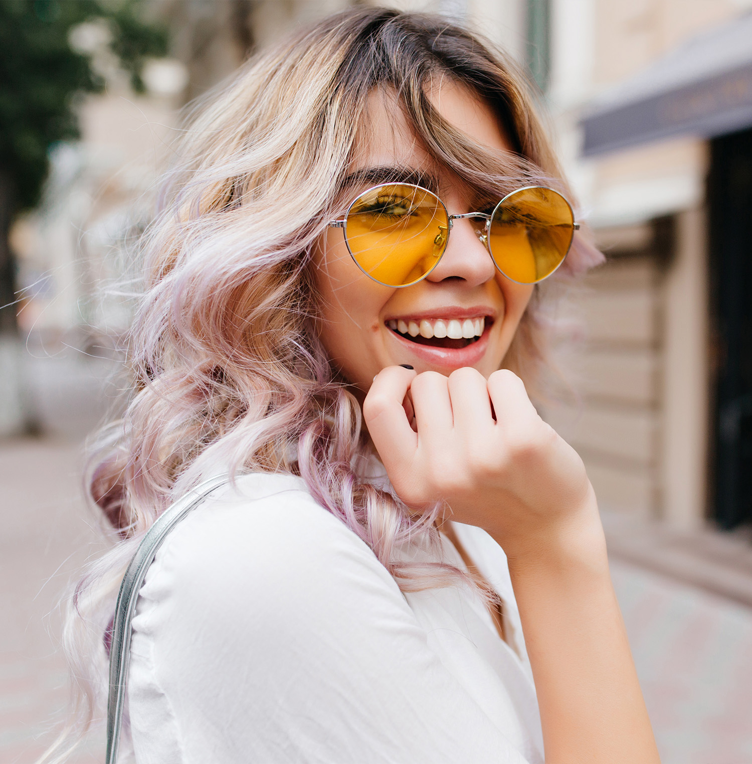 The image shows a woman with long, wavy hair and purple highlights standing outdoors. She is wearing sunglasses with a yellow tint, a white top, and has her hand on her chin with a smile. Behind her is a city street scene with buildings and trees.