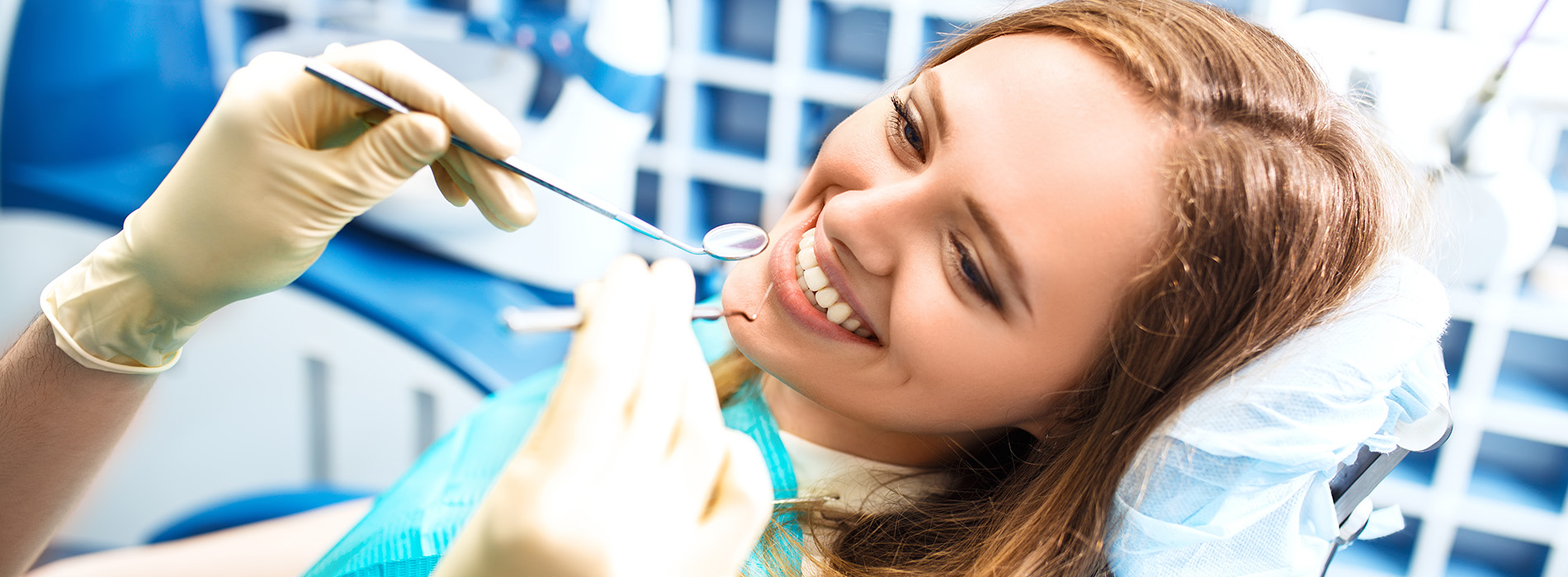 A young woman is sitting in a dental chair, smiling at the camera while getting her teeth cleaned by a professional.