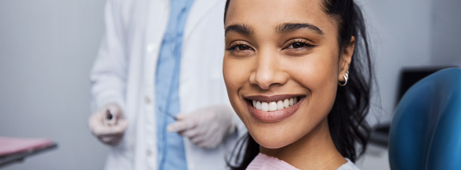 An image of a smiling woman in a dental office, with a dentist in the background.