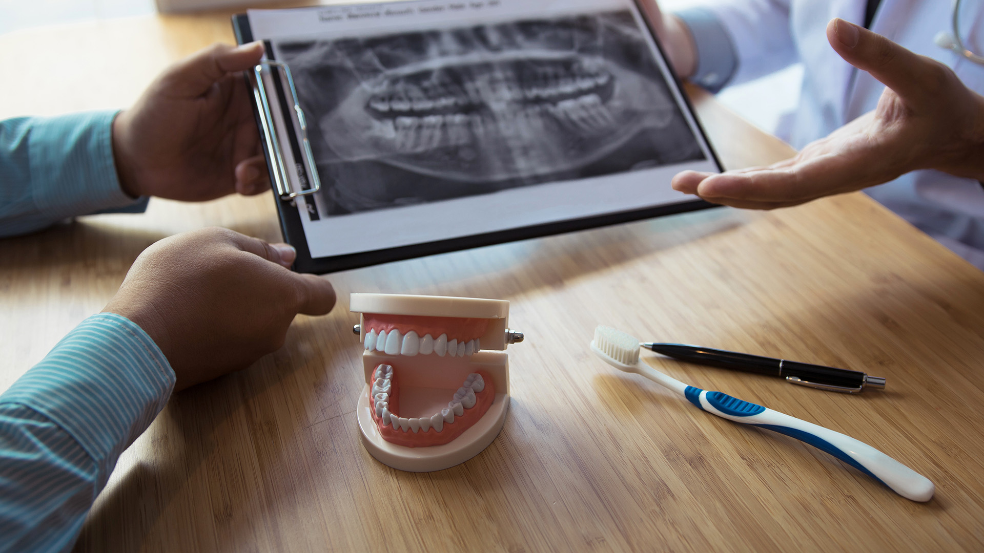 The image depicts a dental professional holding up an X-ray to show a patient, with various dental tools and models visible on the table.