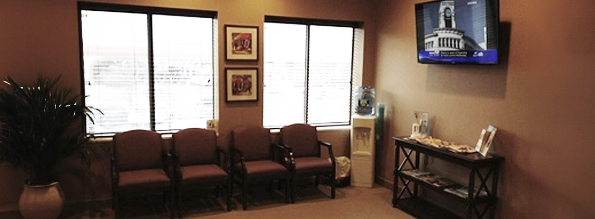 The image shows an interior view of a waiting room in a medical or dental office, with a television displaying news, comfortable seating arrangements, and a framed certificate on the wall.