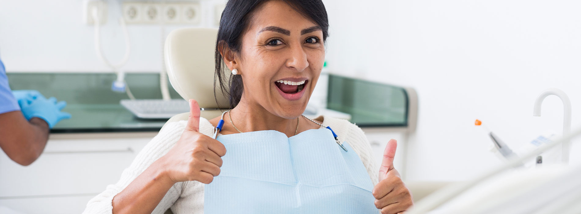 A woman, presumably a dental professional, is smiling and giving a thumbs up while sitting in front of a dental chair.