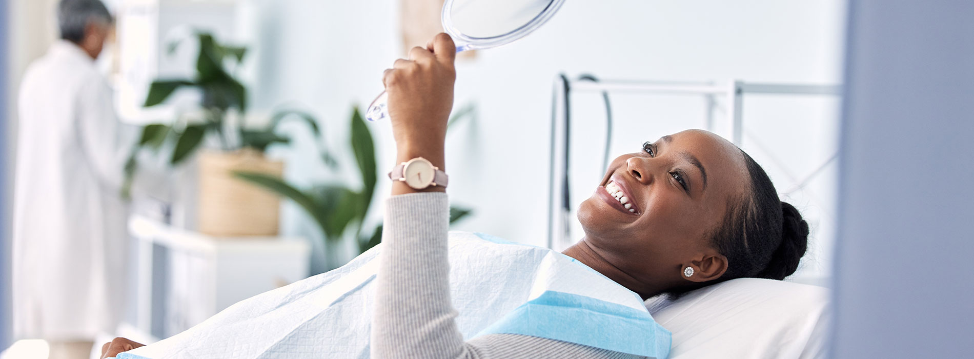 A woman in a hospital bed, smiling and holding up a mirror to her face.