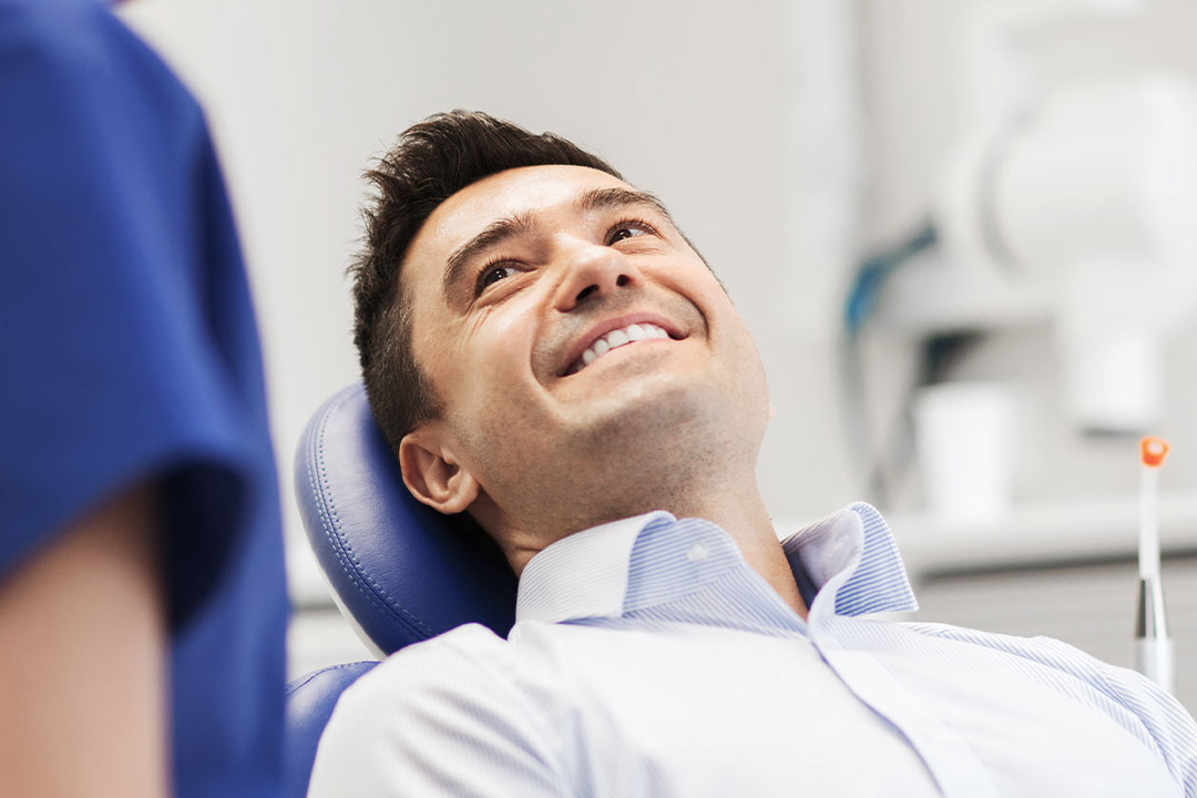 A man is seated in a dental chair, smiling at the camera, with a dental professional standing beside him.