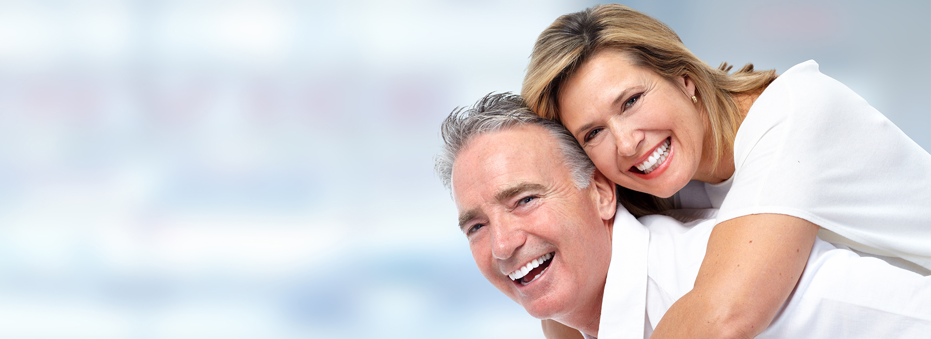 The image is a photograph of an elderly couple embracing and smiling at the camera, with a blurred background that suggests they are indoors.