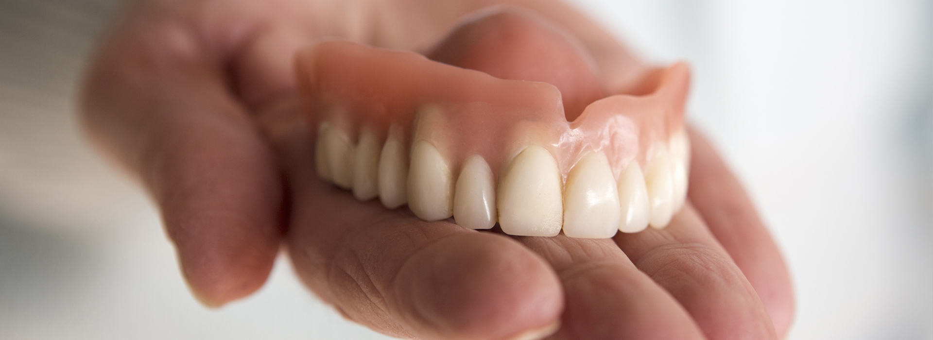 The image shows a hand holding a set of dentures against a blurred background, with the focus on the prosthetic teeth.