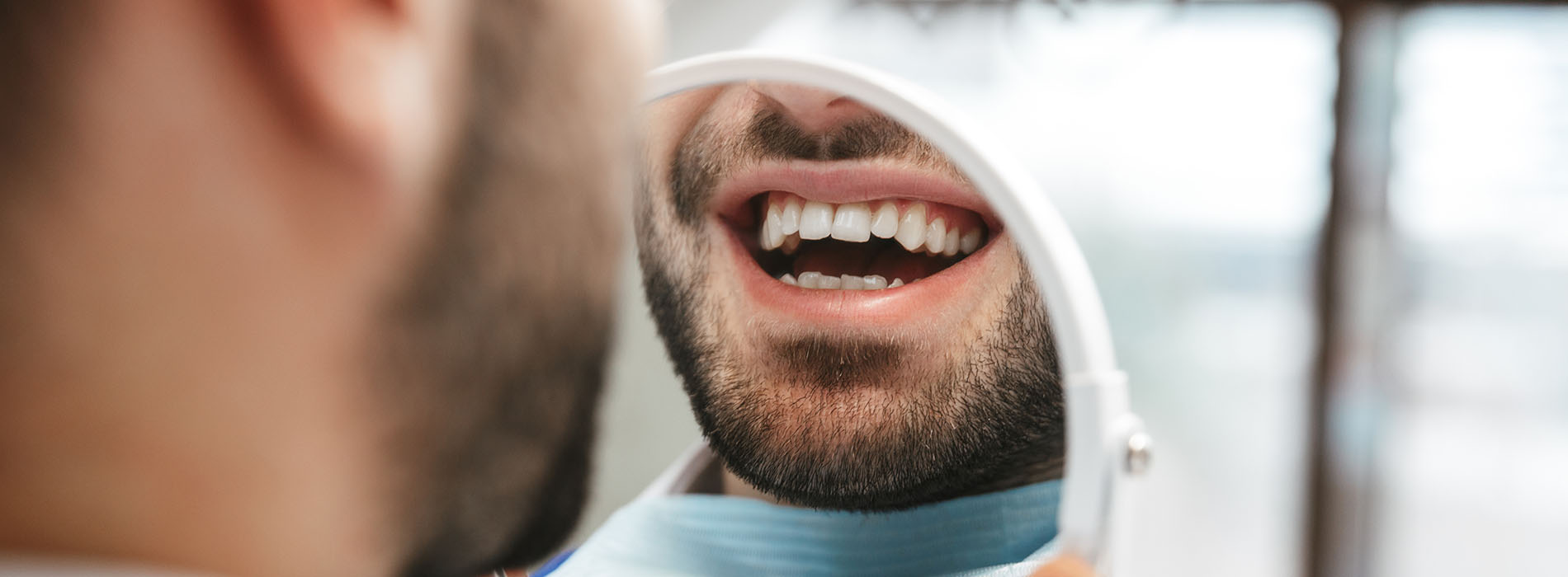 The image shows a man in the foreground with his mouth open, revealing teeth and a tongue piercing. In the background, there is a partial view of another person who appears to be wearing a blue shirt and possibly a surgical mask or face shield.