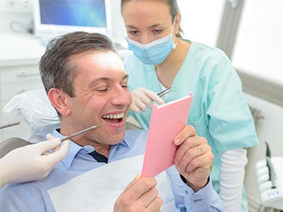 Man sitting in dental chair, smiling broadly while holding up pink card. Dental professional standing behind him, looking at the card with a slight smile.