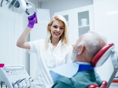 A dental hygienist is assisting an elderly man in a dental chair, with the woman holding a dental mirror and smiling at the camera.