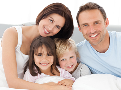 A family of four, including a man, woman, and two children, posing for a photo in bed with smiles.