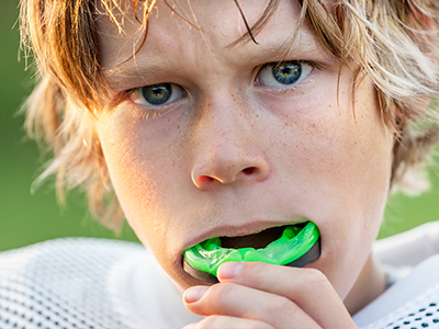 A young male athlete with blonde hair, holding a sports ball in his mouth, and wearing a football uniform.