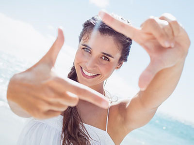 A smiling woman with long hair, standing on a beach, holding up her index finger in front of the camera.