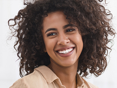 A woman with curly hair is smiling at the camera, wearing a beige top.